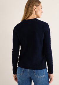 Marinblå tröja med en strukturerad stickdesign, prydd med kabelmönster längs ärmarna och en ribbad nederkant, bärs tillsammans med blå jeans.
