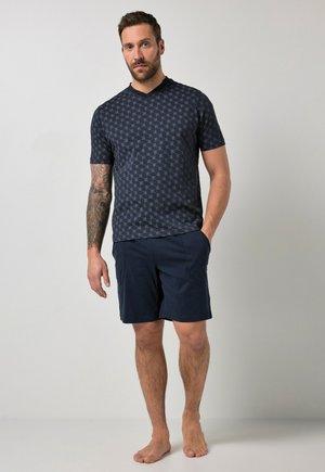 Mand med skæg og tatoveret arm iført en mønstret navy kortærmet skjorte og navy shorts, stående barfodet mod en ensfarvet baggrund.