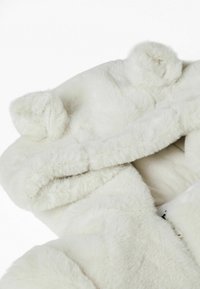 Chaquetón de pelo sintético blanco con textura suave, orejas redondas en la capucha y un diseño acogedor. El interior cuenta con un forro de tela suave.
