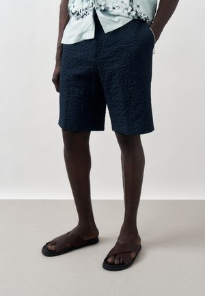 Férfi texturált tengerészkék rövidnadrágban, világoszöld virágos ingben és barna papucsban, zsebében tartott kézzel áll sima padlón.