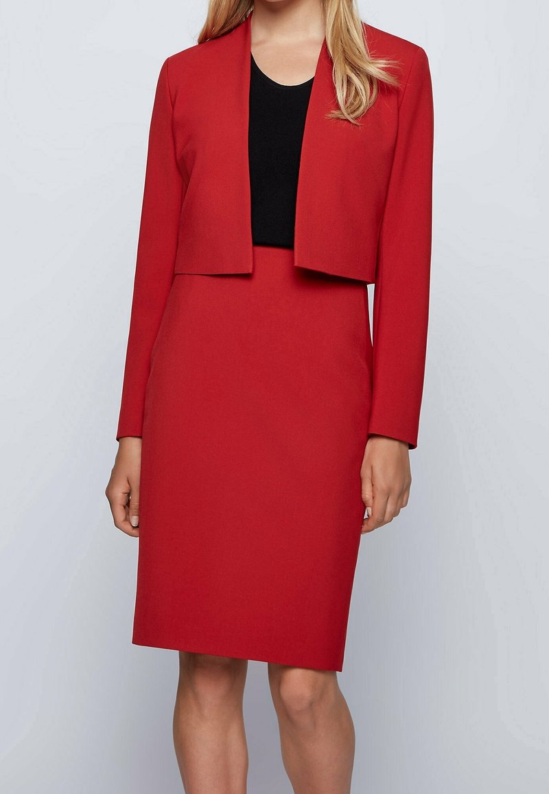 Femme portant un tailleur jupe rouge ajusté avec une veste courte par-dessus un top noir, debout devant un fond uni gris clair.