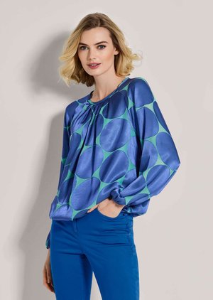 Femme blonde portant un chemisier bleu et vert à gros pois avec manches longues et pantalon bleu, debout avec une main dans la poche.