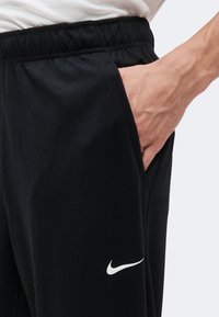 Personne portant un pantalon noir Nike, une chemise blanche, main dans la poche du pantalon, logo Nike swoosh visible près de la poche, taille élastique.