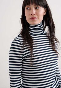 Žena s dugom tamnom kosom koja nosi crno-bijelu prugastu turtleneck vestu, blago gleda lijevo u odnosu na jednostavnu svijetlu pozadinu.