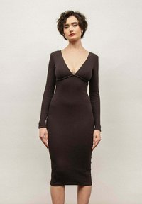 Długa, dopasowana sukienka z długim rękawem w ciemnym brązie, z głębokim dekoltem w kształcie litery V i gładką, elegancką teksturą. Kończy się poniżej kolana.