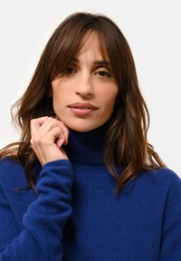 Kobieta z długimi brązowymi włosami, ubrana w niebieski sweter z golfem, trzymająca kołnierz blisko podbródka, patrząca prosto przed siebie.