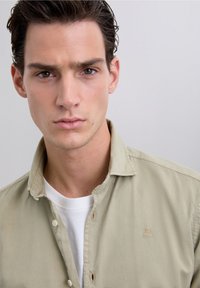 Vaaleanvihreä puuvillainen nappipaita, jossa on rintalogo. Päällä valkoinen pyöreäkauluksinen t-paita. Sileä pinta, rento istuvuus.