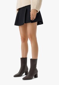 Botas de cuero marrón hasta el tobillo con un diseño estructurado, punta afilada, cremalleras laterales y tacones gruesos, combinadas con una falda negra plisada.