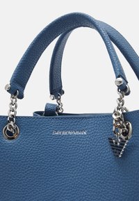 Modrá kožená kabelka s hrubou texturou, stříbrnými akcenty, řetízkovými detaily a logem. Krátké ucha a strukturovaný design jsou výrazné.