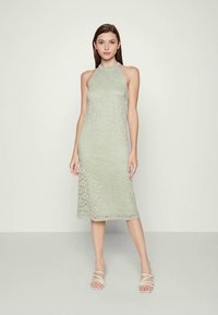 Vestuário midi de renda verde-claro com decote halter, corpete justo e saia em A, combinado com sandálias transparentes de tiras.