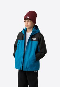 Blauwe en zwarte waterdichte jas met een ritssluiting aan de voorkant en een capuchon. Gedragen met een wit T-shirt en zwarte broek. Heeft een logo op de borst en een beanie.