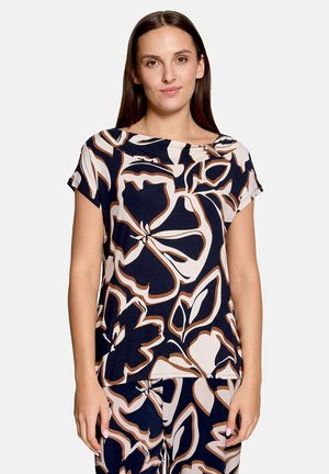 Femme portant un haut à manches courtes et un pantalon assortis avec de grands motifs floraux abstraits bleu marine, blanc et brun, debout devant un fond uni.