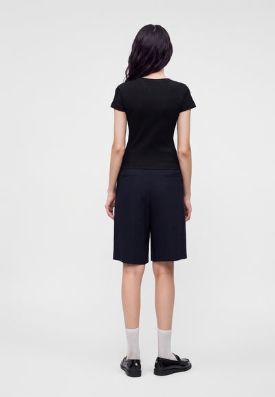 Femme aux longs cheveux foncés porte un haut noir à manches courtes, un short sombre au genou, des chaussettes blanches et des mocassins noirs, debout, de dos.