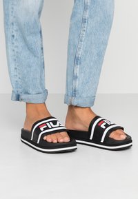 Sandalias de slide negras con acentos y logotipo blancos, suela gruesa blanca, diseño de punta abierta, material suave, combinadas con jeans de denim azul claro enrollados.