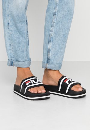 Sandalias de slide negras con acentos y logotipo blancos, suela gruesa blanca, diseño de punta abierta, material suave, combinadas con jeans de denim azul claro enrollados.