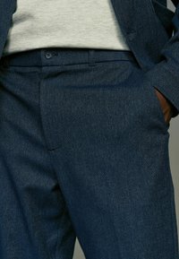 Pantaloni sartoriali blu scuro realizzati in tessuto texturizzato, con una tasca frontale e una chiusura a bottone singolo in vita.