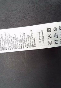 Vit tvättlapp i tyg med svart text. Innehåller tvättsymboler, materialinformation (100% ull) och flera språköversättningar.