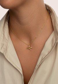 Collana d'oro con ciondolo a forma di piccolo aereo, catena delicata con perline, posizionata su un colletto di camicia di colore neutro.