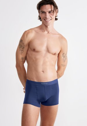 Uśmiechnięty mężczyzna bez koszulki, ubrany w granatowe bokserki marki Sloggi, stojący na tle jednolitego jasnego tła z tatuażami na obu ramionach.