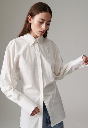 Femme aux longs cheveux bruns, portant une chemise blanche ample boutonnée et un jean bleu, regardant vers le bas avec un bras légèrement levé.