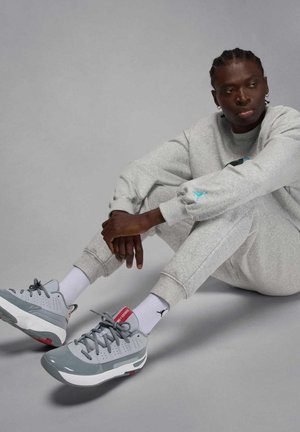 Mężczyzna z zaplecionymi warkoczami, ubrany w szary dres i szare sneakersy z białymi skarpetkami, siedzi na podłodze na szarym tle.