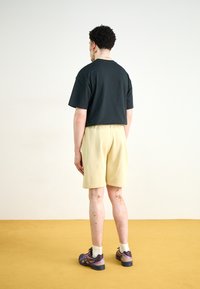 Svart t-skjorte med korte ermer, lysegule shorts, svarte og lilla joggesko, vist fra baksiden på et gult gulv.