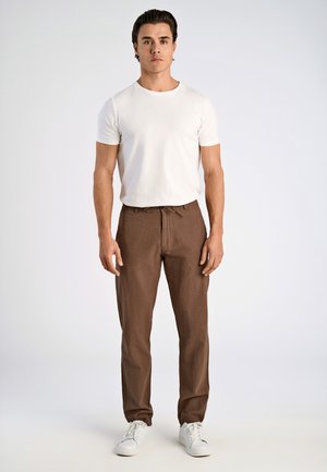 Una camiseta blanca de manga corta combinada con pantalones sueltos de color marrón, texturizados y con cintura elástica. El modelo lleva zapatillas blancas.