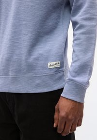 Εγκαρτέρης ενός ατόμου που φοράει ένα ανοιχτό μπλε υφασμάτινο πουλόβερ με μικρή ετικέτα "Scotch & Soda" κοντά στην άκρη και μαύρο παντελόνι.