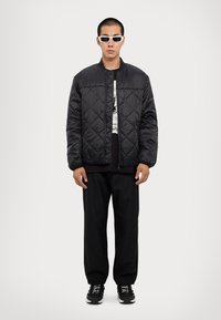 Padded black bomber jacket with diamond pattern, zip closure, elastic cuffs, layered over a black graphic t-shirt and black sweatpants.