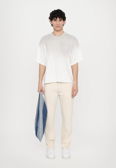 White oversized t-shirt with a raw neckline, paired with light beige slim-fit pants and white sneakers. Holding a blue and gray patterned fabric.