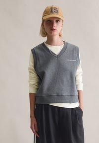 Graue Weste mit V-Ausschnitt, über einem hellgelben Langarmshirt getragen. Kombiniert mit dunkler Hose; mit kleinem Logo auf der Brust.