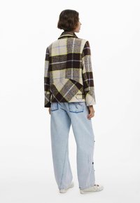 Gehäkelte Wolljacke mit braunen, gelben und blauen Mustern, ausgestattet mit einem Reverskragen und lockerer Passform, kombiniert mit hellen Jeanshosen.