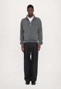 Sudadera de forro polar gris con cremallera de media longitud y texto bordado, combinada con pantalones cargo negros de pierna ancha y zapatos negros. Textura suave.
