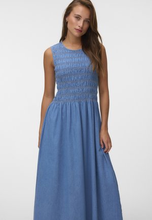 Femme aux longs cheveux bruns portant une robe sans manches bleu clair avec un corsage froncé et une jupe fluide sur un fond uni.