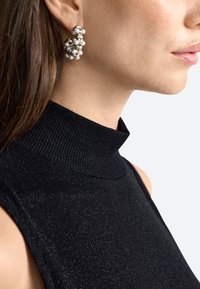 Zwart geribbeld high-neck top gecombineerd met een opvallende multi-parel oorring met zilveren hardware, vastgelegd vanuit zijaanzicht.