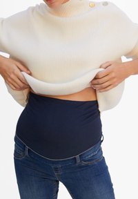 Suéter blanco de canalé con un cuello alto y dos botones, llevado sobre una prenda ajustada de color azul marino y jeans azules.