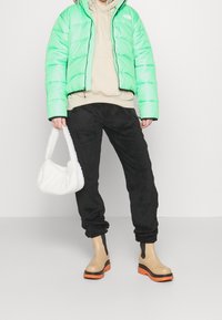 Person iført en lysegrøn puffjakke, beige hoodie, sorte fuzzy bukser, beige støvler med orange såler, og holder en hvid fluffy håndtaske.