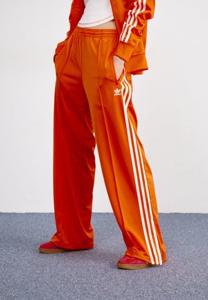 Persona con pantalones deportivos Adidas naranjas y chaqueta a juego con rayas blancas, combinados con zapatos rojos, de pie sobre una alfombra gris.