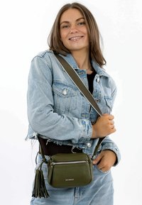 Olive green leather crossbody bag with a textured finish, featuring a zip closure and tassel accent. Worn with a denim jacket.