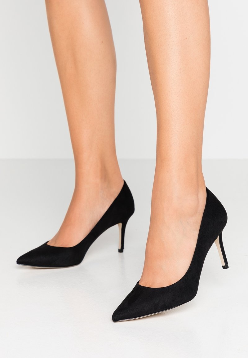 Les escarpins en daim noir présentent un bout pointu et un talon stiletto. L'empeigne a une texture lisse avec une ouverture de cheville sans couture.