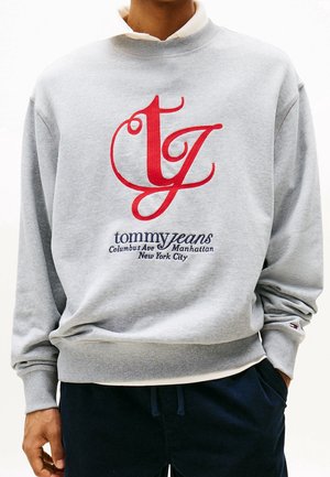 Persoon draagt een lichtgrijze trui met een rood "tg"-logo en de tekst "tommy jeans Columbus Ave Manhattan New York City" aan de voorkant.