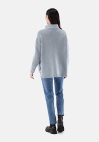 Hellgrauer gestrickter Pullover mit hohem Kragen, oversized Passform und seitlichen Schlitzen. Kombiniert mit blauen Jeans und schwarzen klobigen Stiefeln.