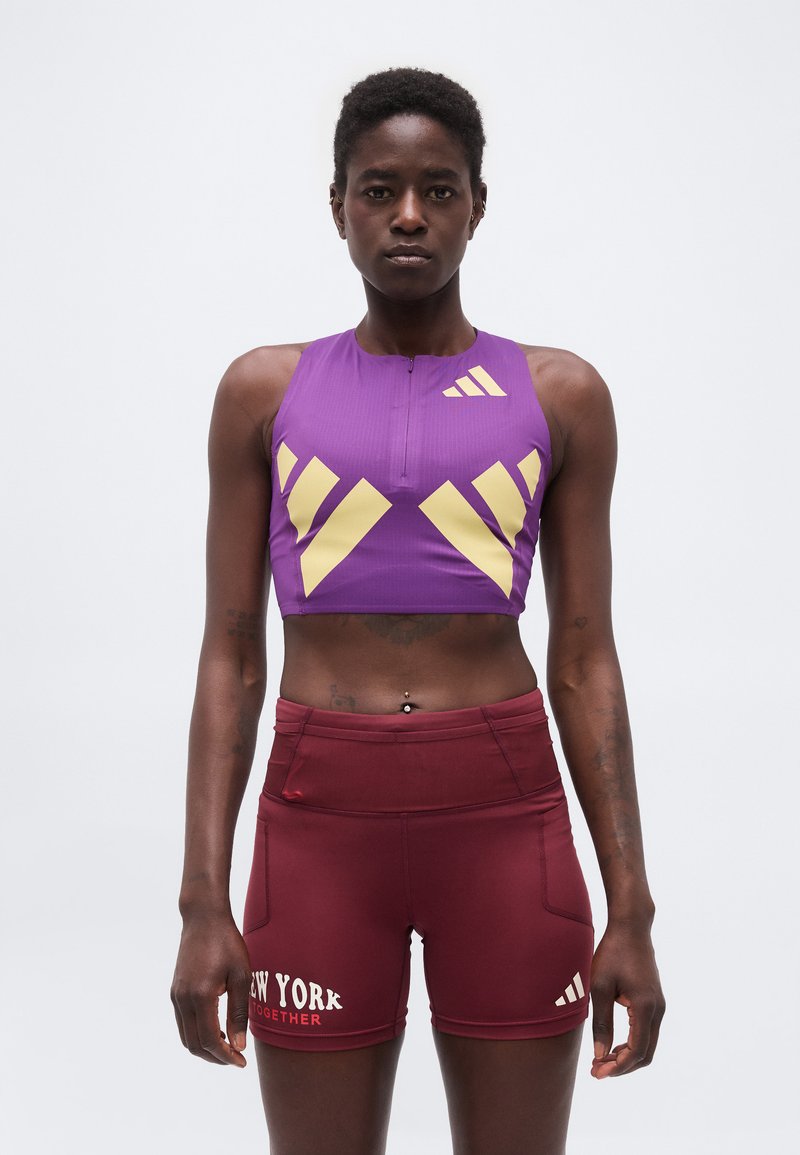 Αθλήτρια με μοβ και κίτρινο crop top και μπορντό σορτς με το κείμενο «New York Together», στέκεται μπροστά από ένα απλό φόντο.