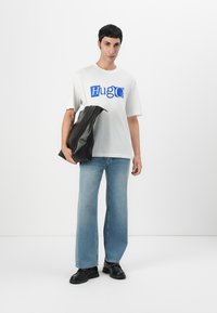 Camiseta blanca con logo azul "Hugo", combinada con jeans de pierna ancha de un azul claro y zapatos de cuero negros. El modelo sostiene un bolso negro.