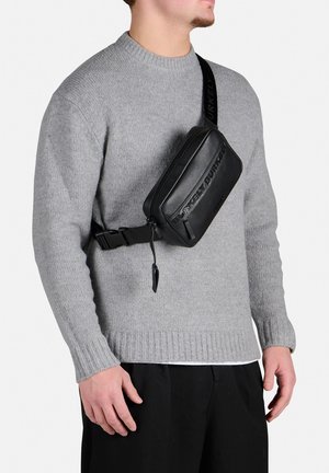 Borsa a tracolla in pelle nera con logo impresso, chiusura con zip e tracolla regolabile. Indossata sopra un maglione grigio, sono visibili contrasti di texture.