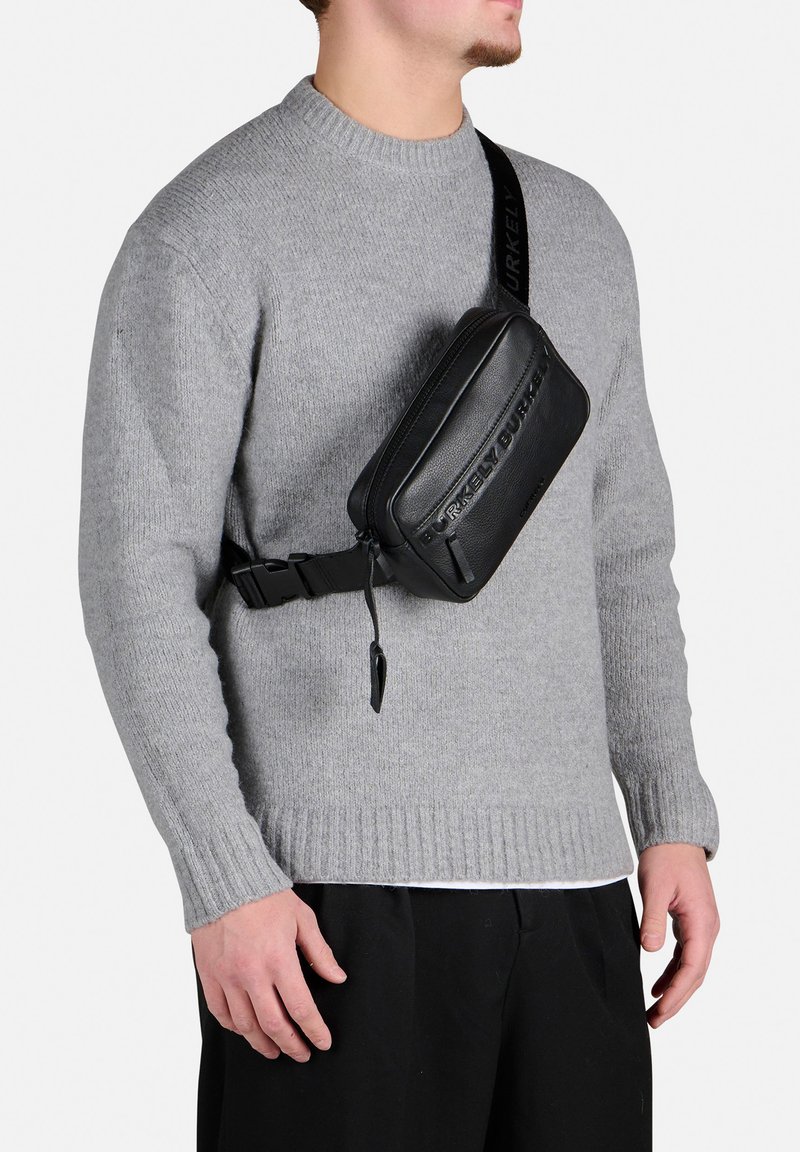 Borsa a tracolla in pelle nera con logo impresso, chiusura con zip e tracolla regolabile. Indossata sopra un maglione grigio, sono visibili contrasti di texture.