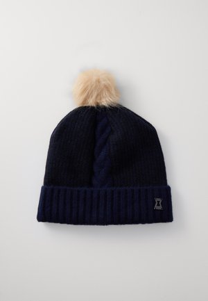 Mornarica pletena kapa z ribastim robom in vzorcem cable knit, s krznjeno pom-pom na vrhu in majhnim logotipom na strani.
