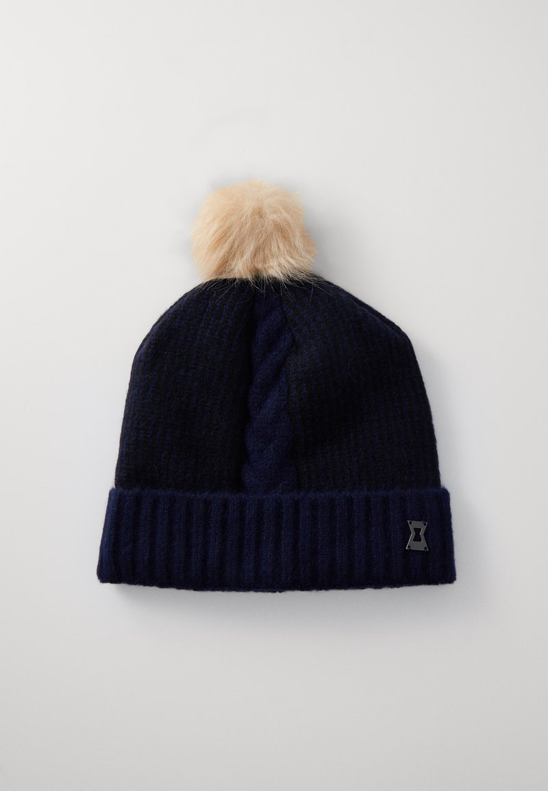 Marineblå strikket lue med ribbestrikket kant og kabelstrikket mønster, med en pelsdusk på toppen og en liten logo-detall på siden.