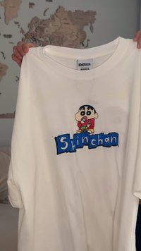 Bílé bavlněné tričko s kreslenou postavičkou s černými vlasy a červeným tričkem, pod kterou jsou modrými blokovými písmeny napsána slova "Shinchan".