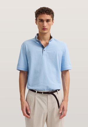 Junger Mann trägt ein hellblaues Polo mit kurzen Ärmeln, beige Hose und einen dunklen geflochtenen Gürtel, steht vor neutralem Hintergrund.